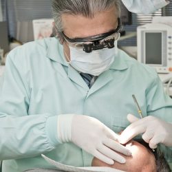 Dentista con bata verde, gafas de aumento y mascarilla examinando a un paciente